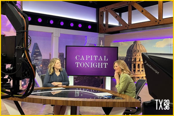 Two women sit in a TV studio at desk in front of a TV screen that has the show title, "Capital Tonight," on it. There are two large studio cameras in the foreground.