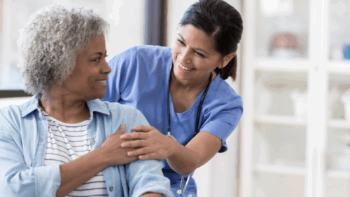 A seated woman with grey hair wearing a blue shirt smiling and touching hands with a standing, smiling nurse wearing blue scrubs and a stethoscope around her neck.